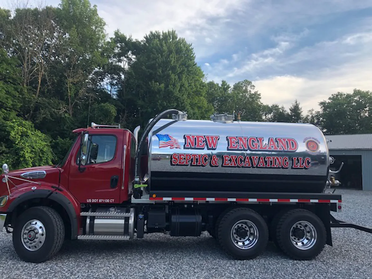 The company's red Freightliner truck with a polished stainless steel septic tank, the company name hand lettered on the tank in red beside a US flag, parked on gravel at the edge of the woods
